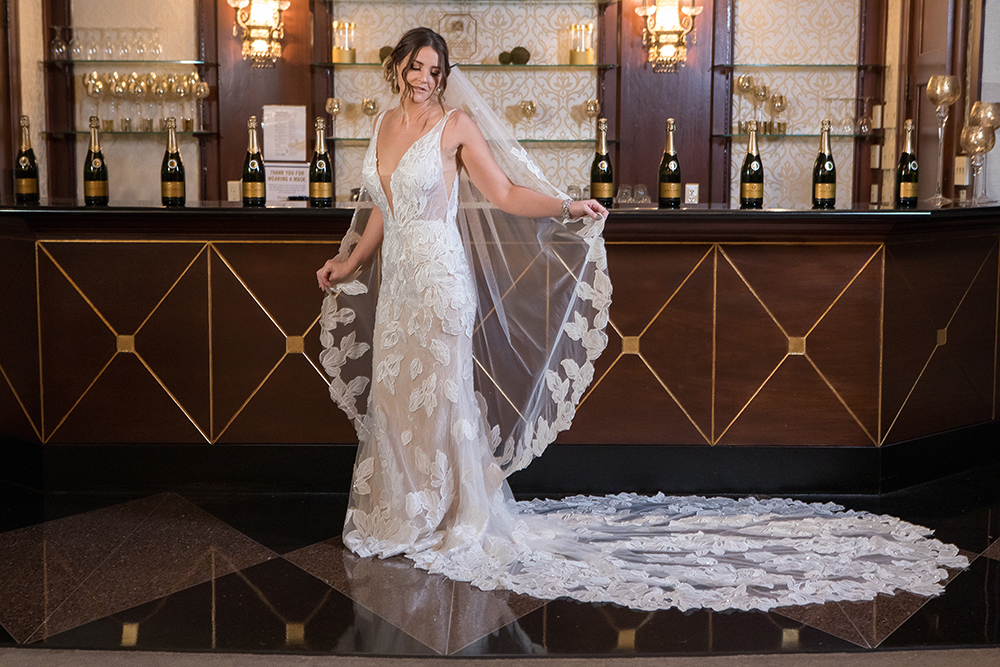 Evie is wearing an ivory lace fit and flare gown with deep V-plunge and dramatic cascading train available at MaeMe The Bridal Boutique. Matching veil available. For her “Something Blue” Passport Polish’s short length Ready Made Press-ons in one of their newest colors, Iceland.