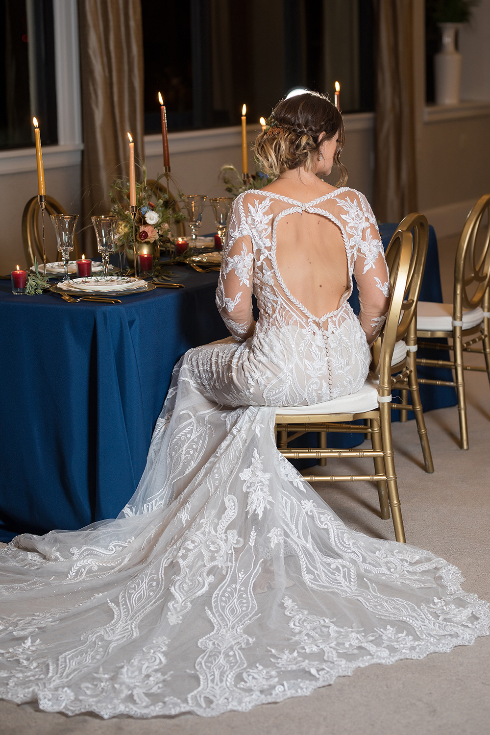 Evie is wearing a graphic lace fit-and-flare wedding dress with illusion sleeves from MaeMe The Bridal Boutique. Scrolling patterns and ornate structures shape and sculpt the figure, from the narrowed plunge of the V-neckline to the sheer cutouts along the sides and back. A clasp a the top of the back creates a wide keyhole opening, framed with organic floating laces to complete this head-turning look.