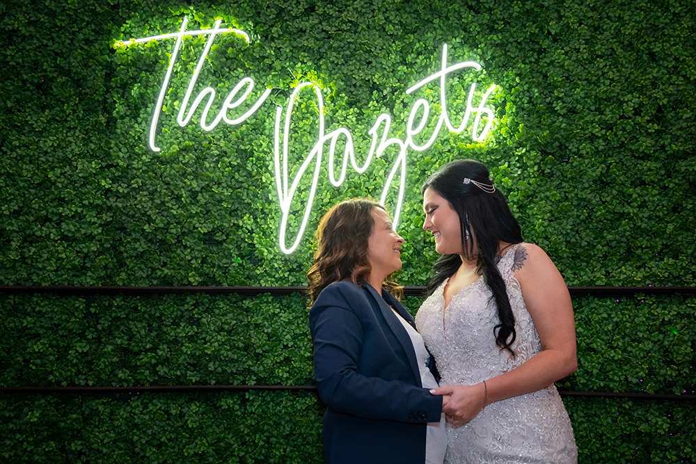 the brides pose in front of a greenery wall with custom neon sign